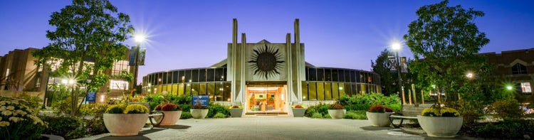 Modern building with a sun emblem above the entrance, lit at night with bright lights, trees, and planters.